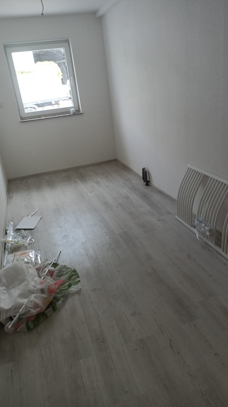 Pusty pokój z ułożonymi panelami podłogowymi w kolorze jasnego drewna, widoczne śmieci po remoncie, grzejnik żeberkowy oraz okno