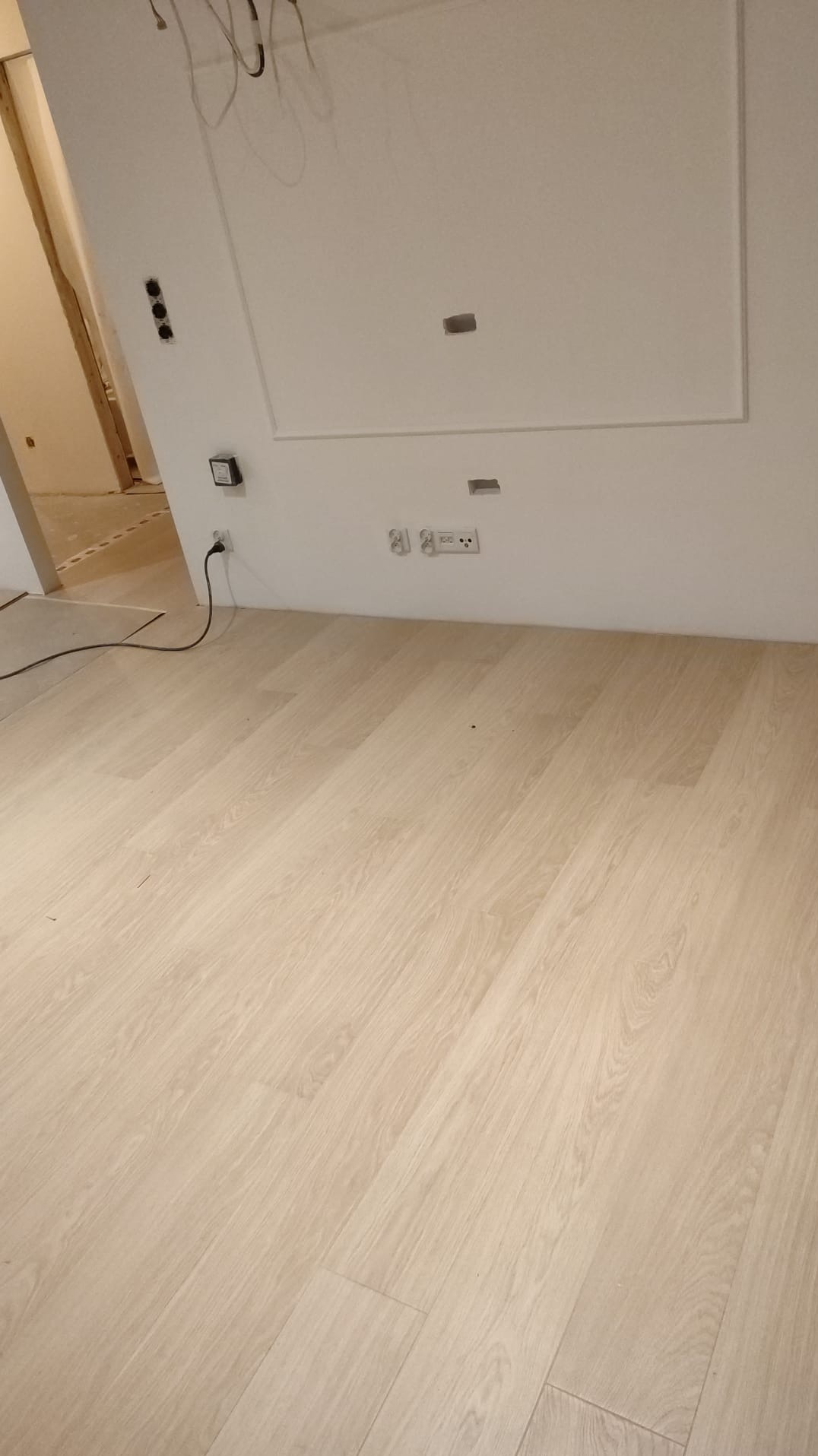 Jasne panele podłogowe ułożone w nowym mieszkaniu, widoczne gniazdka elektryczne i przygotowane otwory w ścianie.