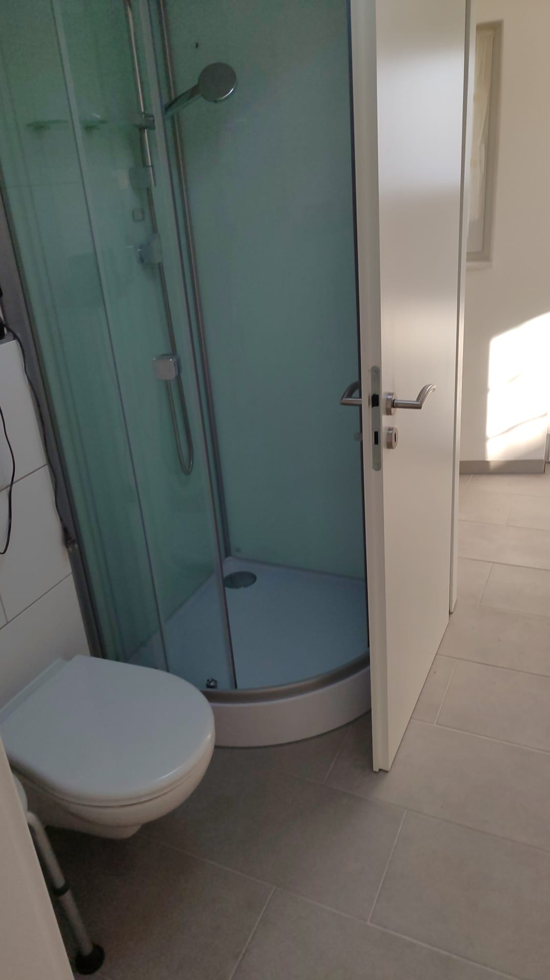 Nowoczesna kabina prysznicowa z okrągłym brodzikiem i szklanymi drzwiami, widok z otwartych drzwi do łazienki, jasne płytki podłogowe, toaleta w rogu.