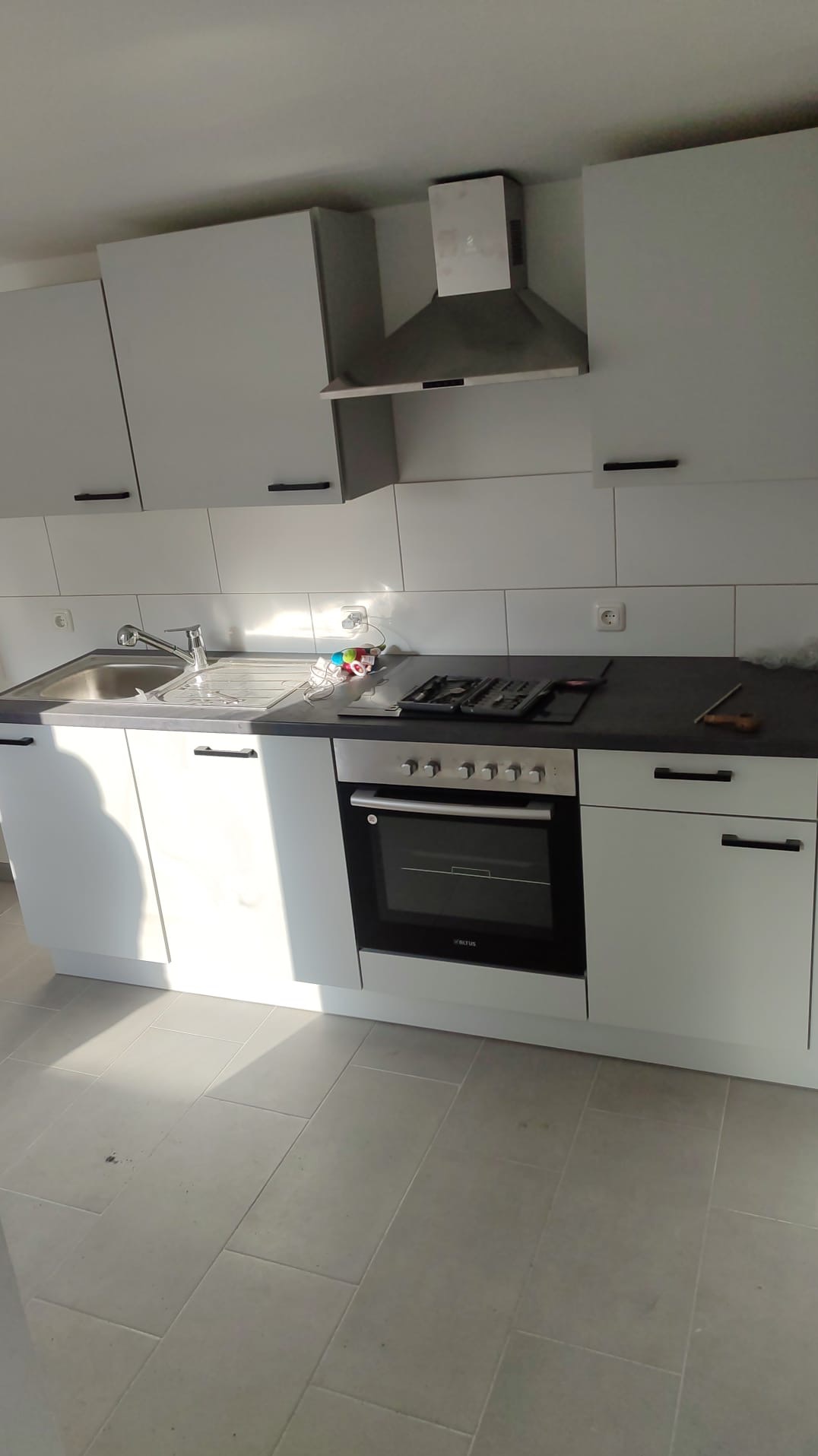 Jasna, nowoczesna kuchnia z białymi szafkami, ciemnym blatem, płytą gazową i piekarnikiem Altus, wykończona jasnymi płytkami podłogowymi i białymi płytkami ściennymi.