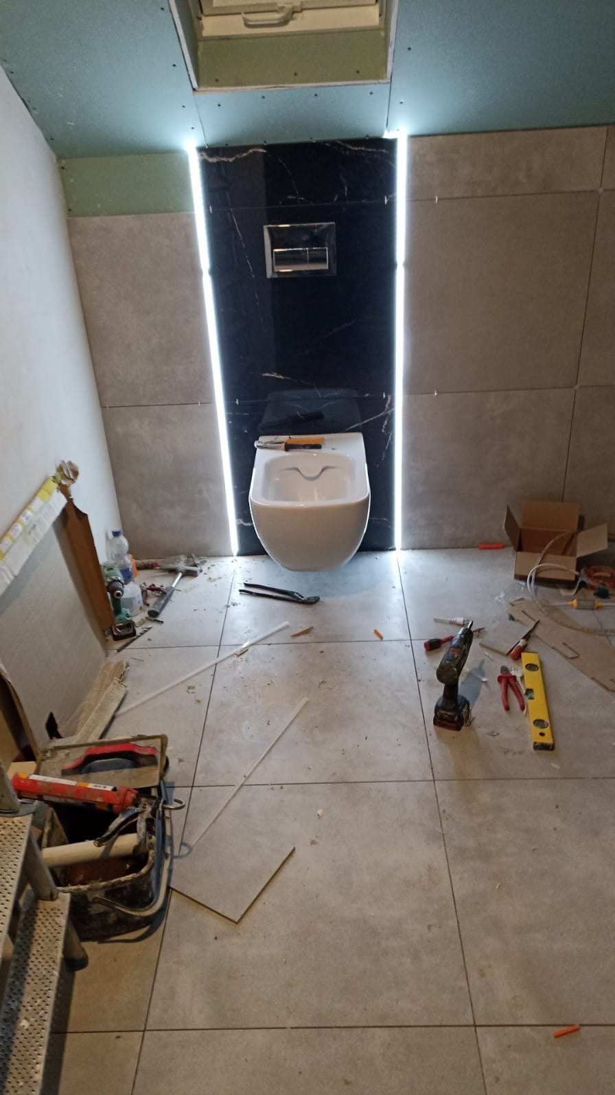 Montaż podwieszanej toalety na tle czarnej płyty z marmurowym wzorem, podświetlonej po bokach, widoczne narzędzia glazurnicze i płyty podłogowe oraz częściowo położona glazura ścienna.