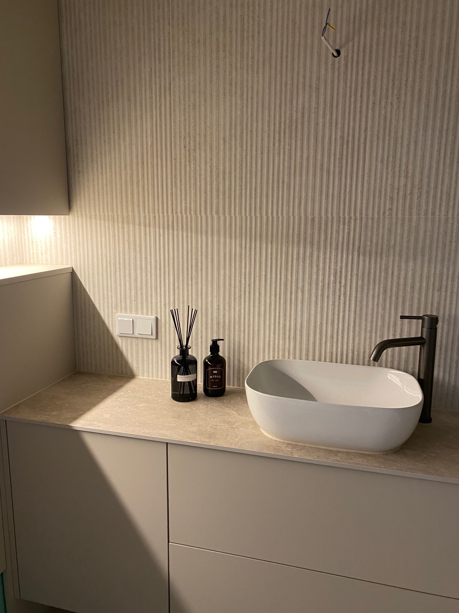 Nowoczesna umywalka nablatowa w kształcie elipsy z czarną baterią, uzupełniona o dyfuzor zapachowy i dozownik mydła, Tomaszów Lubelski. Ściana wyłożona płytkami z delikatnym, pionowym wzorem.