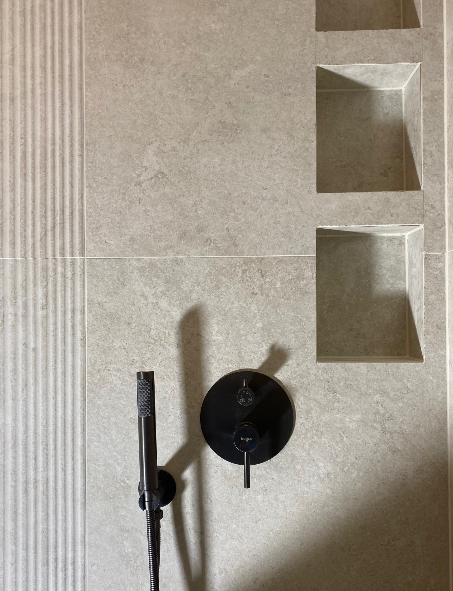 Beżowa ściana z płytek w kabinie prysznicowej, z wnękami i czarną armaturą podtynkową, w tym deszczownicą ręczną i baterią z logo 'begco'.