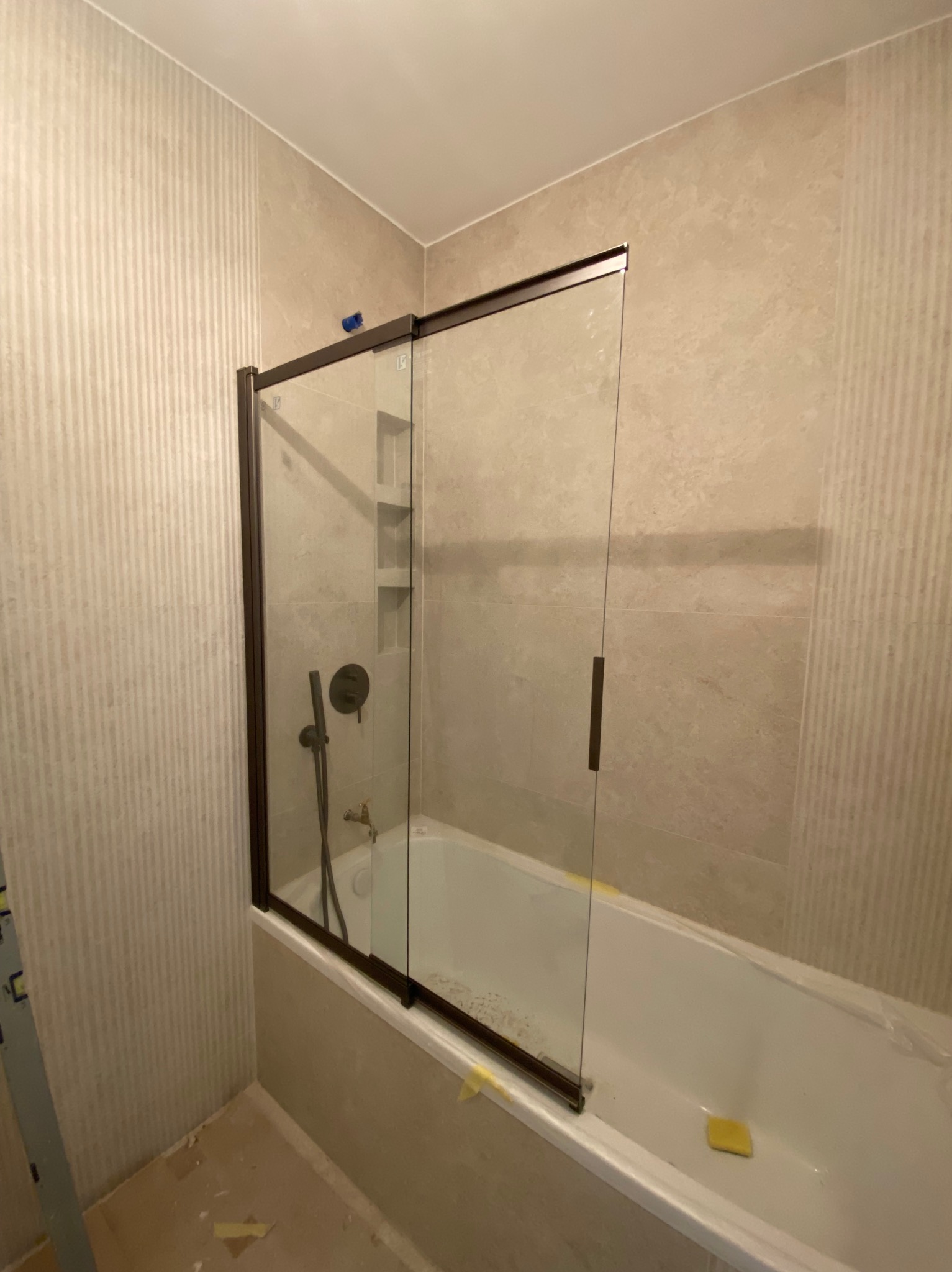 Nowoczesna kabina prysznicowa z przesuwnymi drzwiami w łazience wykończonej beżowymi płytkami, widoczne wnęki na kosmetyki i bateria w kolorze grafitowym.