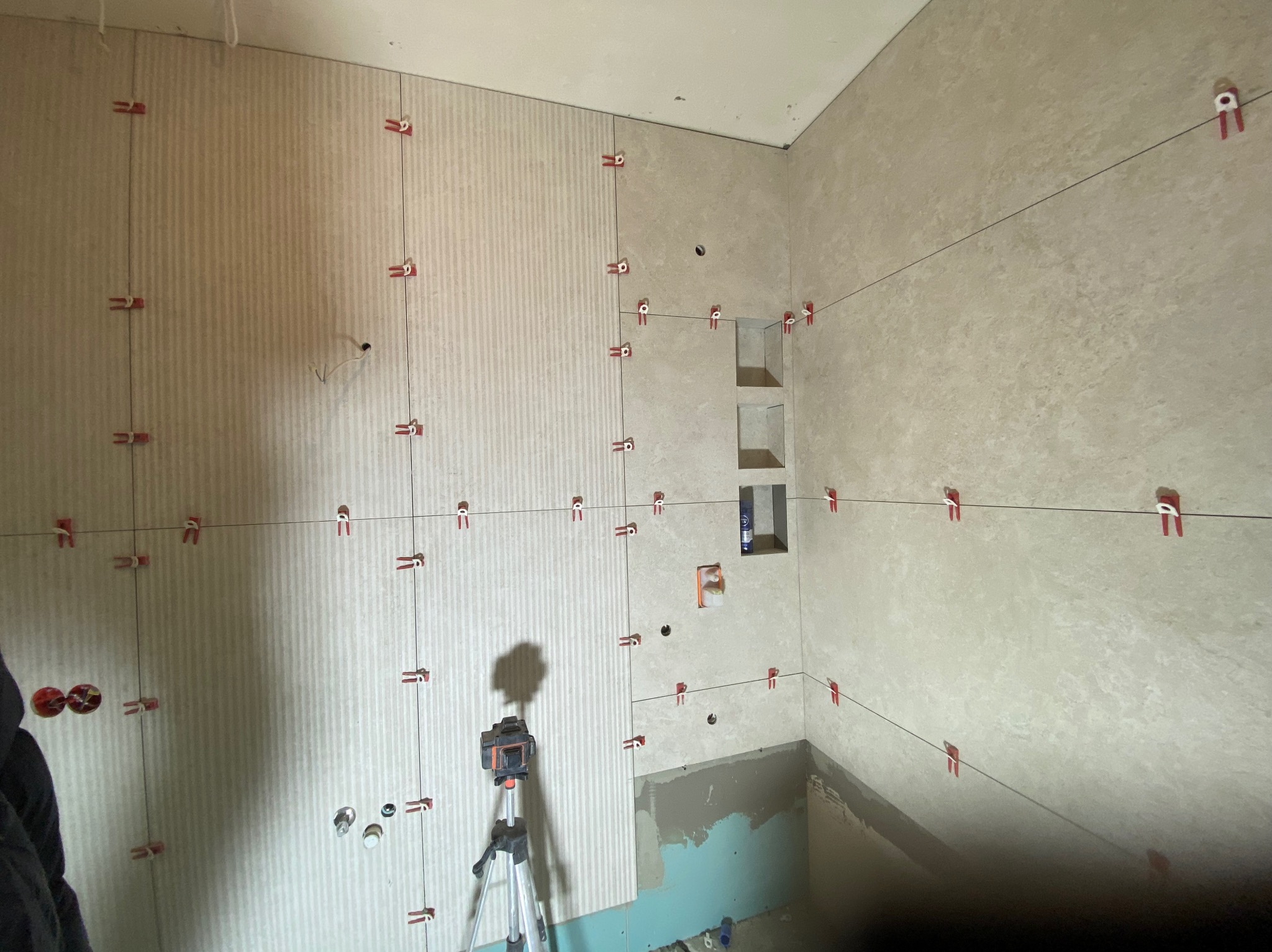 Ściany łazienki w trakcie układania płytek, widoczne poziomice laserowe, krzyżyki dystansowe i częściowo położone płytki w dwóch różnych wzorach.
