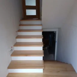 Jasne drewniane schody z białymi podstopnicami prowadzące w dół, widoczna drewniana podłoga i drzwi na górze schodów.