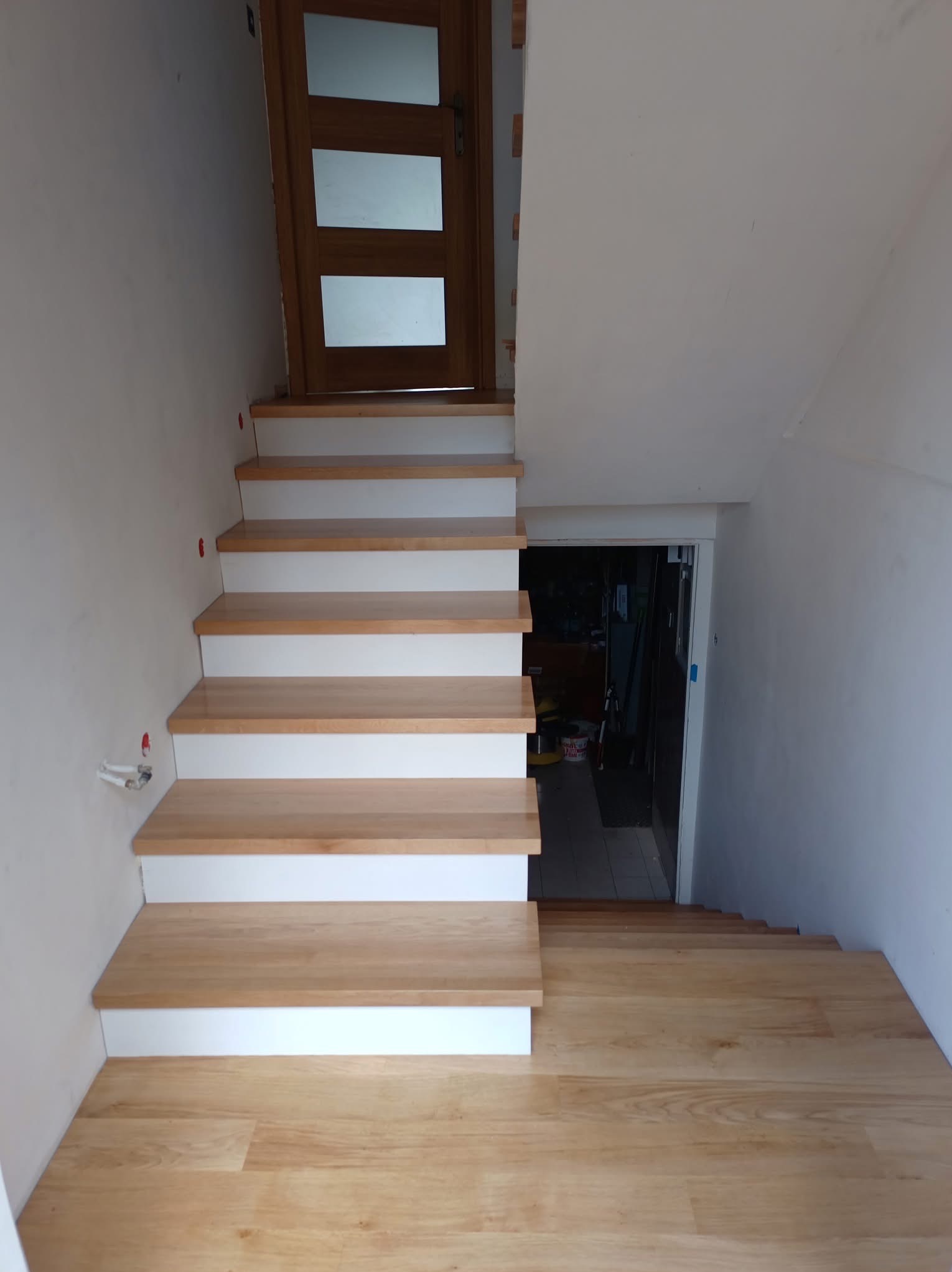 Jasne drewniane schody z białymi podstopnicami prowadzące w dół, widoczna drewniana podłoga i drzwi na górze schodów.