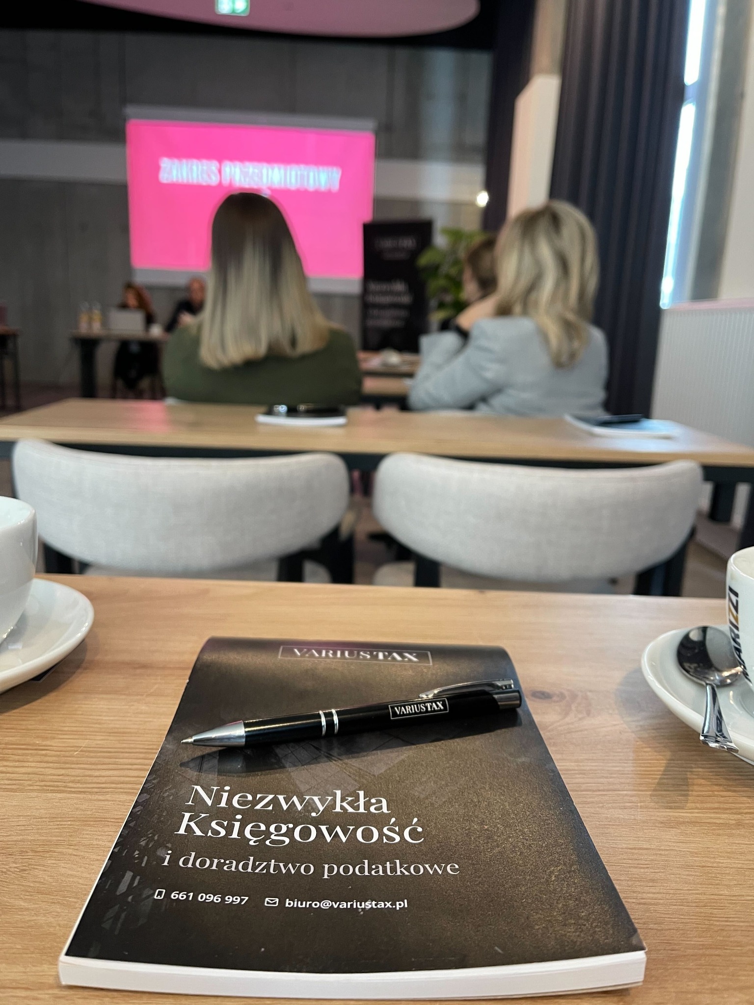 Książka z tytułem 'Niezwykła Księgowość i doradztwo podatkowe' oraz długopisem leżąca na stole w kawiarni, w tle osoby siedzące przy stole i prezentacja.
