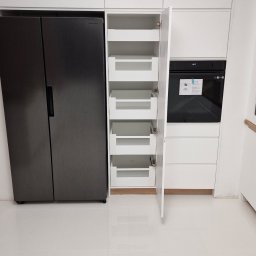 Gorzów - Nowoczesna, biała zabudowa kuchenna z szufladami cargo i piekarnikiem, obok lodówka side-by-side. Minimalistyczny design, połyskliwe fronty, białe płytki podłogowe.