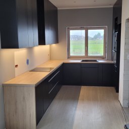 Gorzów - Nowoczesna kuchnia z czarnymi szafkami i drewnianym blatem podświetlonym ledami. Widok na zielone pole przez okno. Minimalistyczny design i funkcjonalność.
