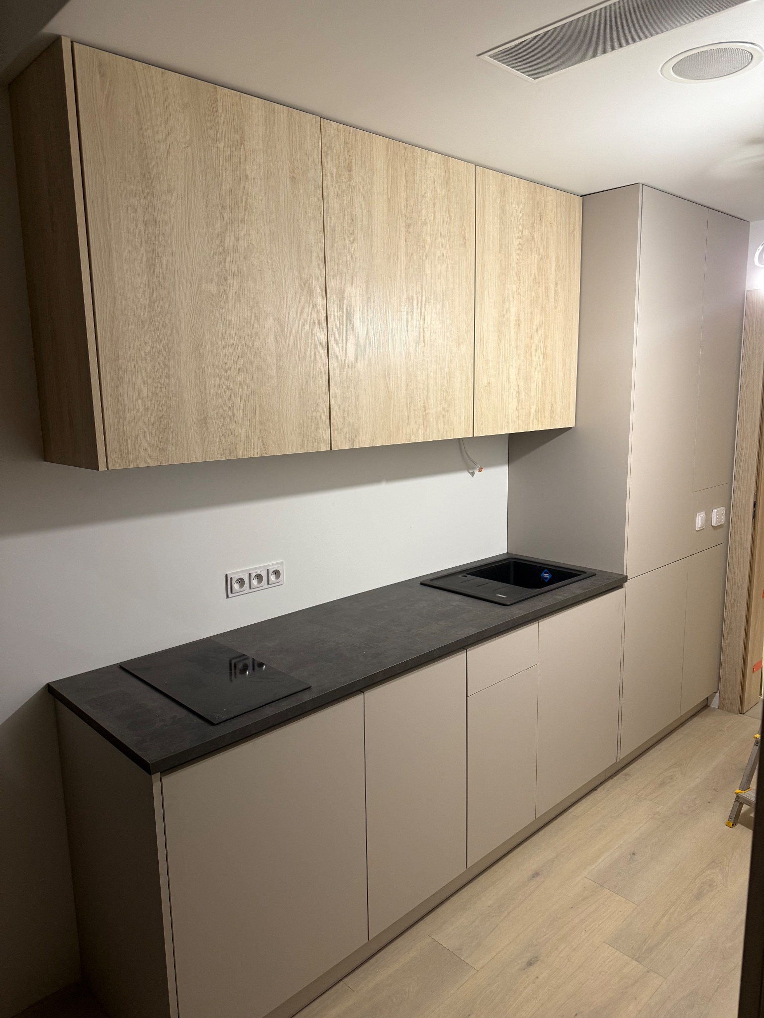 Jasna kuchnia z drewnianymi szafkami wiszącymi i ciemnym blatem. Widoczna płyta indukcyjna i zlewozmywak. Minimalistyczny design, gładkie fronty szafek.