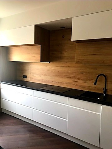 Nowoczesna kuchnia z białymi szafkami i drewnianym panelem ściennym. Czarny blat i bateria dodają elegancji. Minimalistyczny design.
