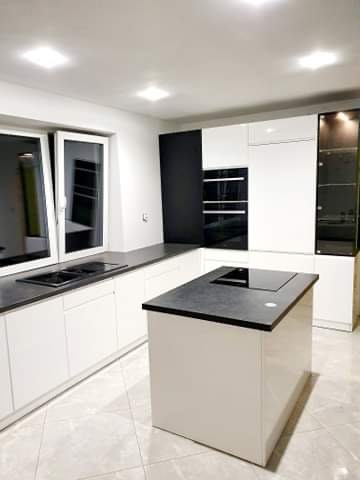 Nowoczesna, jasna kuchnia z wyspą i czarnymi akcentami. Minimalistyczny design, białe fronty szafek, czarny blat i sprzęt AGD. Duże okno wpuszcza naturalne światło.