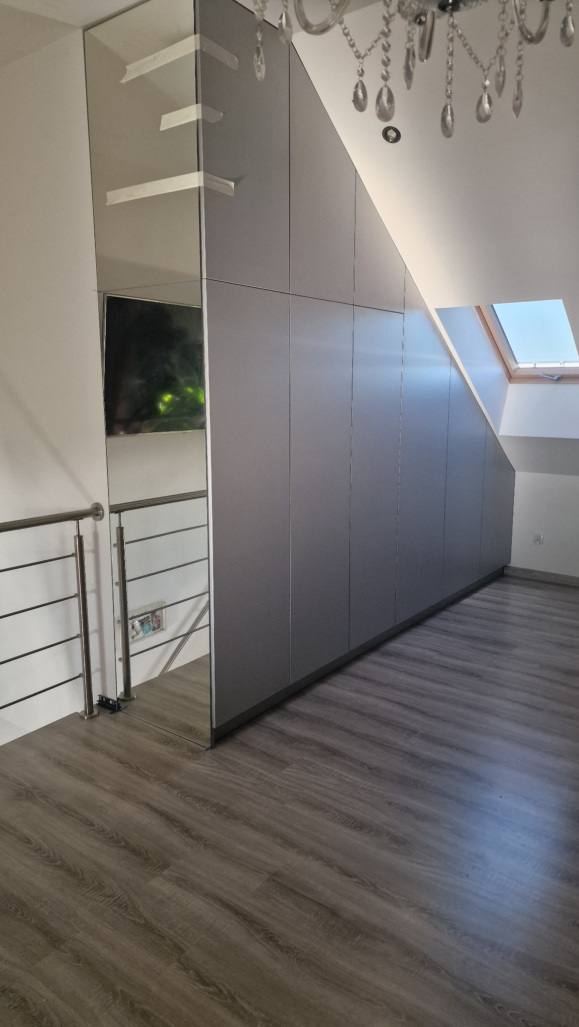 Szafa wnękowa na poddaszu ze szklanym frontem, szara, zabudowa pod skosem, widoczny fragment balustrady i żyrandola, drewniana podłoga.