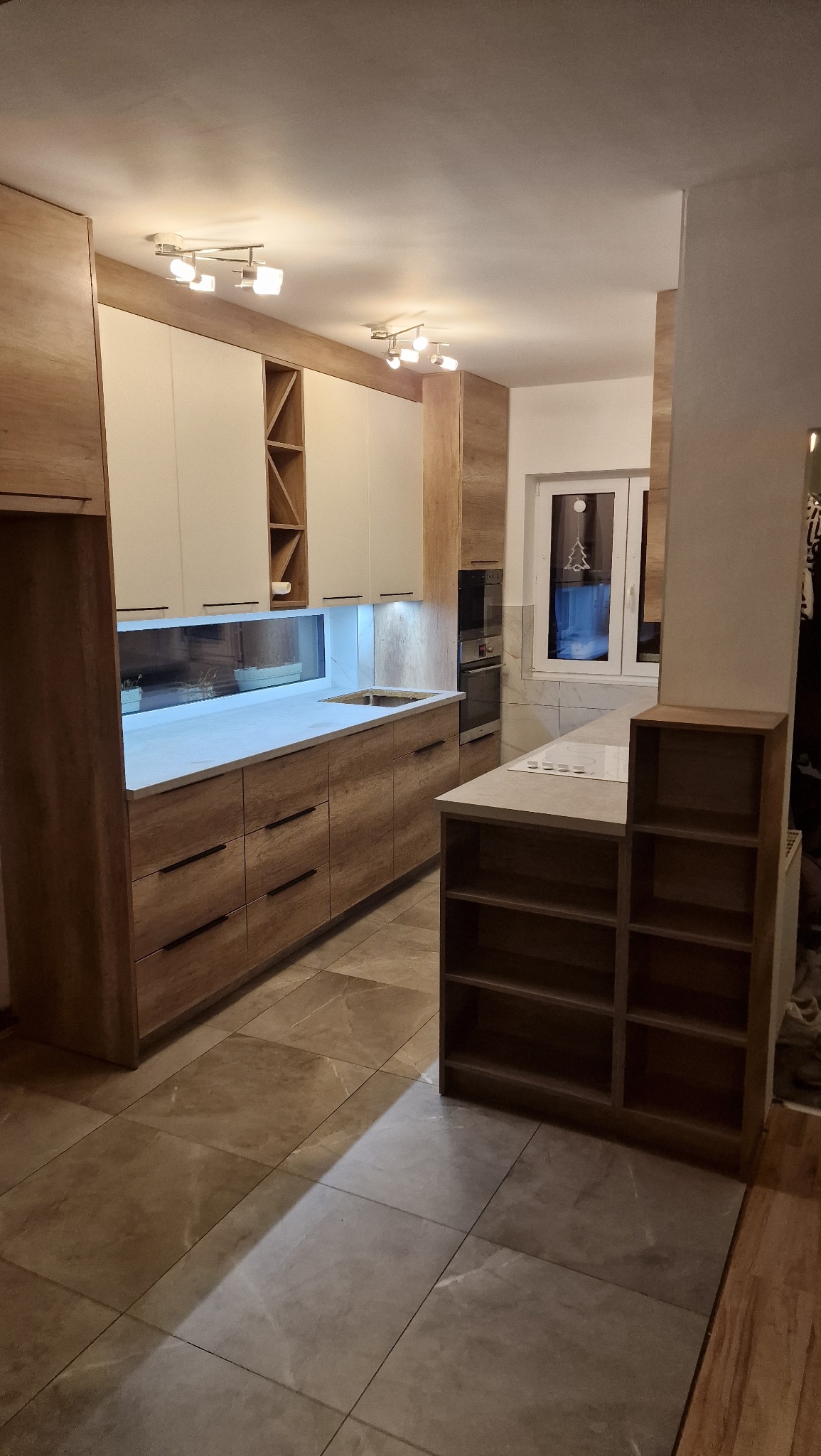 Nowoczesna kuchnia z drewnianymi frontami i białymi szafkami wiszącymi, oświetlona listwą LED pod szafkami. Na blacie widać zlew. W tle okno z dekoracją.