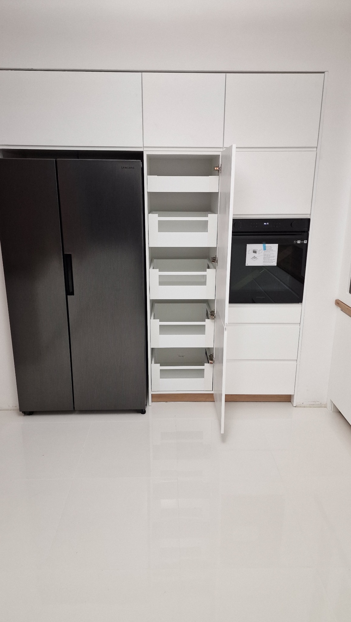 Nowoczesna, biała zabudowa kuchenna z szufladami cargo i piekarnikiem, obok lodówka side-by-side. Minimalistyczny design, połyskliwe fronty, białe płytki podłogowe.