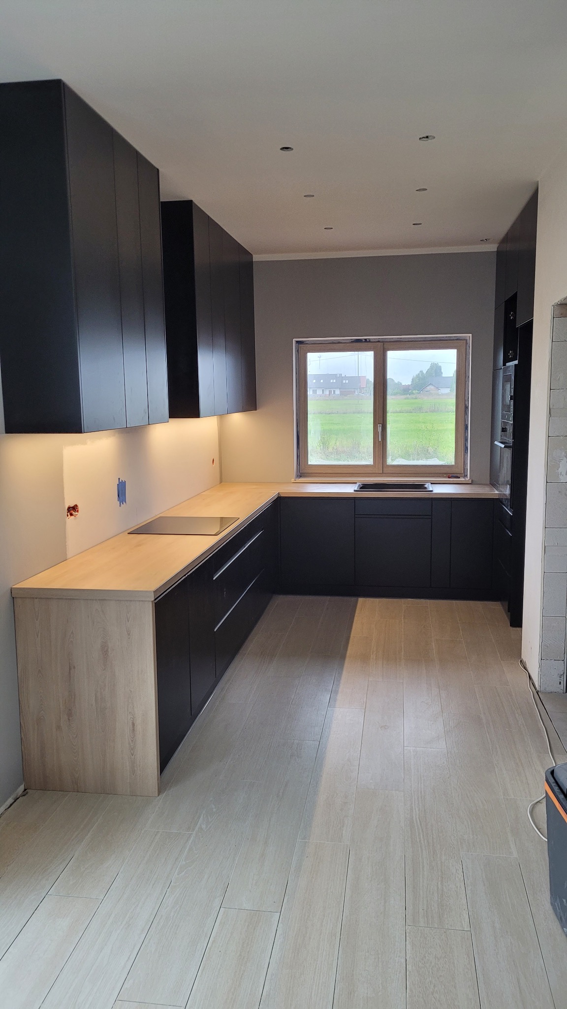 Nowoczesna kuchnia z czarnymi szafkami i drewnianym blatem podświetlonym ledami. Widok na zielone pole przez okno. Minimalistyczny design i funkcjonalność.