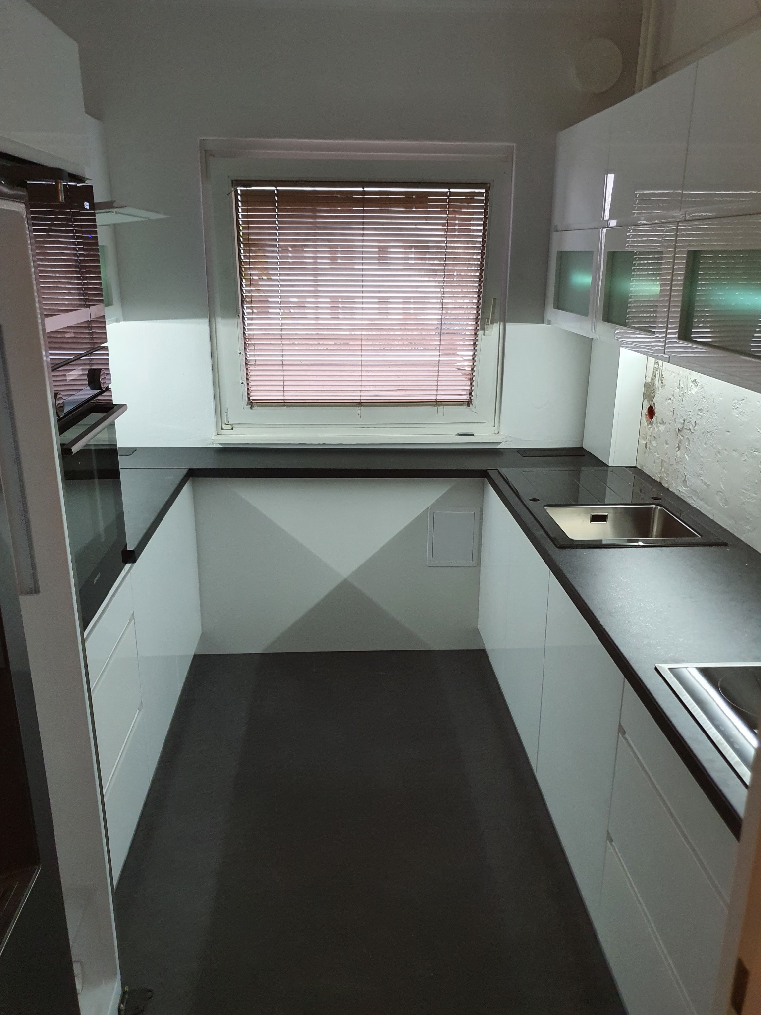 Wąska kuchnia z białymi frontami i ciemnym blatem. Widok na okno z żaluzjami. Minimalistyczny design, oświetlenie LED pod szafkami, nowoczesne wykończenie.