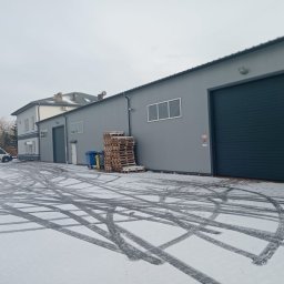 BAJA KRZYSZTOF JAWORSKI - Szary budynek przemysłowy z bramą rolowaną, stertą palet, koszami na śmieci i śladami opon na śniegu, widoczny biały van w tle.