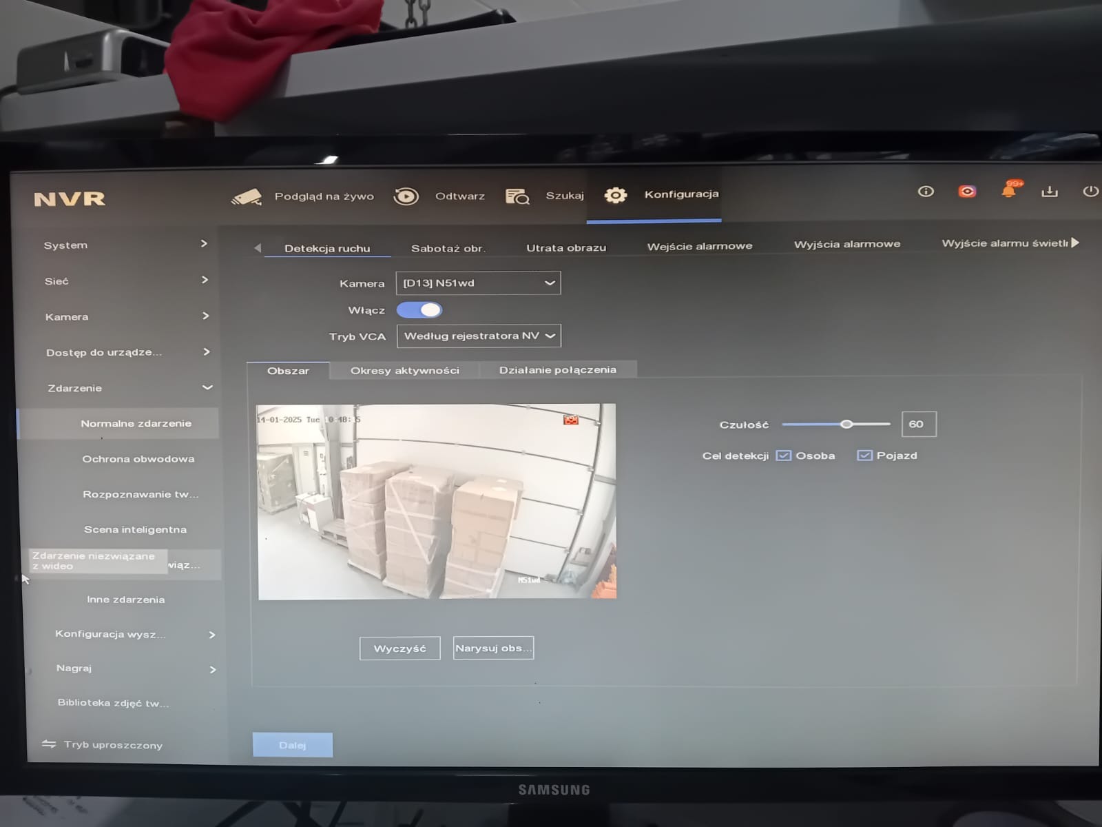 Ekran monitora z interfejsem oprogramowania NVR do monitoringu wizyjnego, wyświetlający obraz z kamery na magazynie z wykrytymi osobami i pojazdami oraz ustawieniami czułości detekcji.