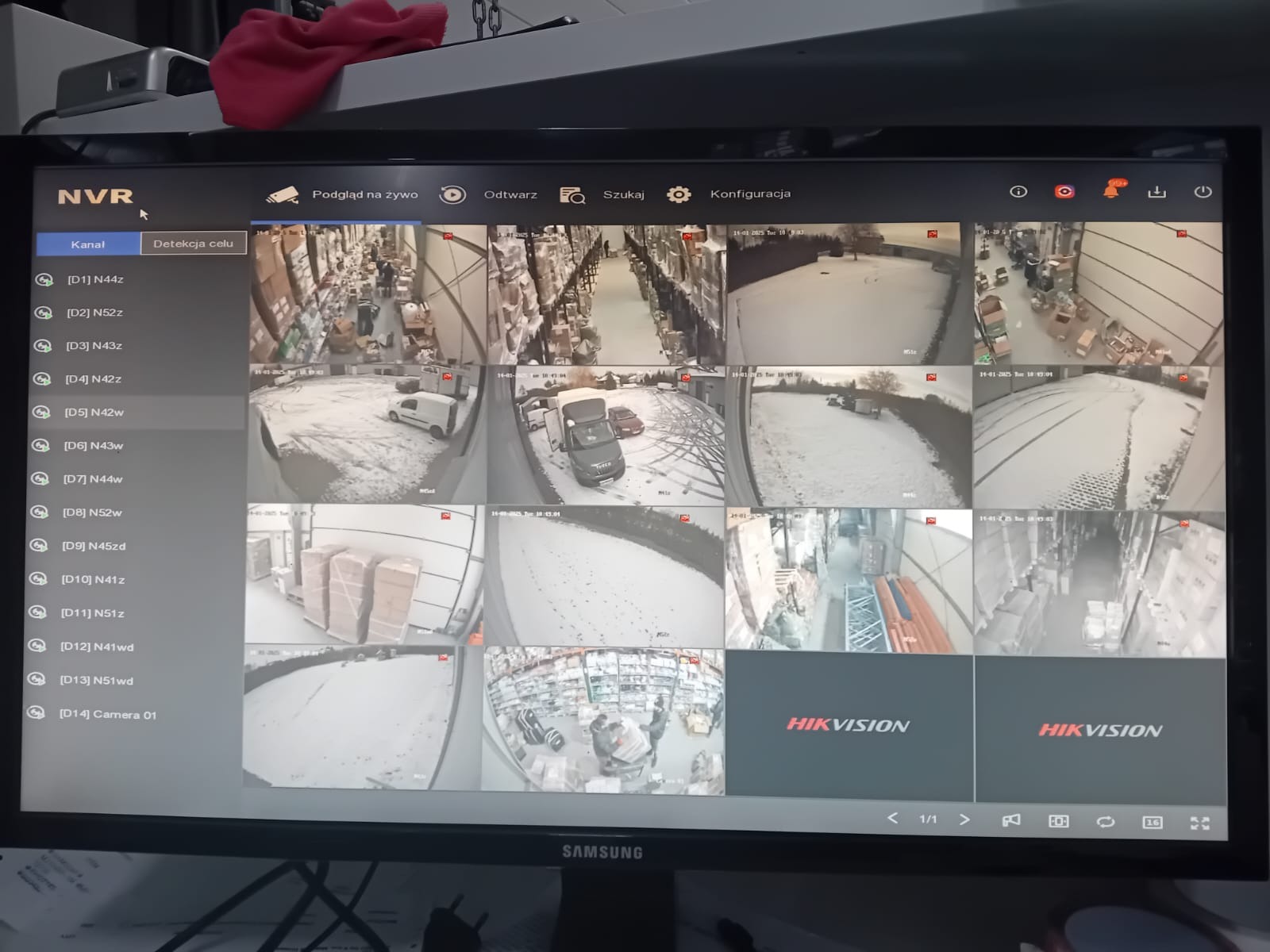 Ekran monitora z podglądem z kamer przemysłowych Hikvision, prezentujący widoki z różnych lokalizacji, w tym magazynu i terenu zewnętrznego pokrytego śniegiem, z interfejsem NVR na górze.