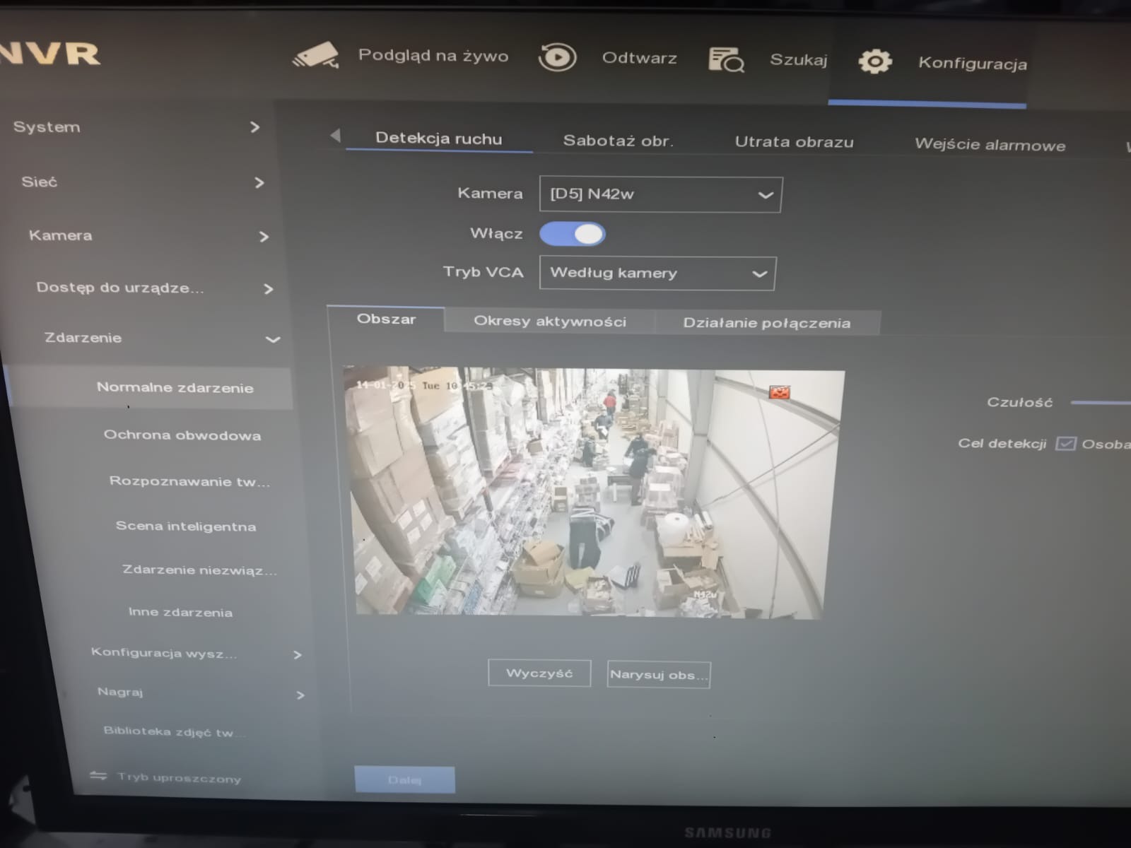 Ekran monitora NVR z ustawieniami detekcji ruchu i podglądem z kamery przemysłowej w magazynie, widoczne osoby i regały z towarem.