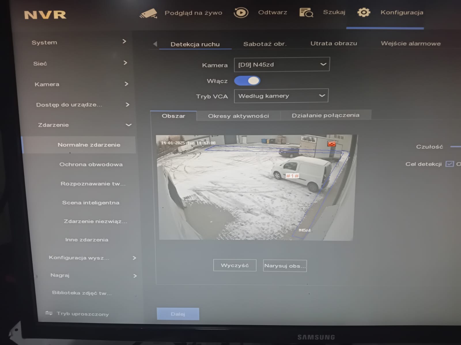 Interfejs oprogramowania NVR z włączoną detekcją ruchu, pokazujący obraz z kamery monitoringu na zewnątrz budynku z widocznym samochodem dostawczym i śladami opon na śniegu.