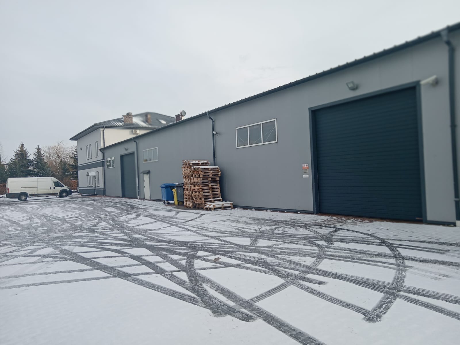 Szary budynek przemysłowy z bramą rolowaną, stertą palet, koszami na śmieci i śladami opon na śniegu, widoczny biały van w tle.
