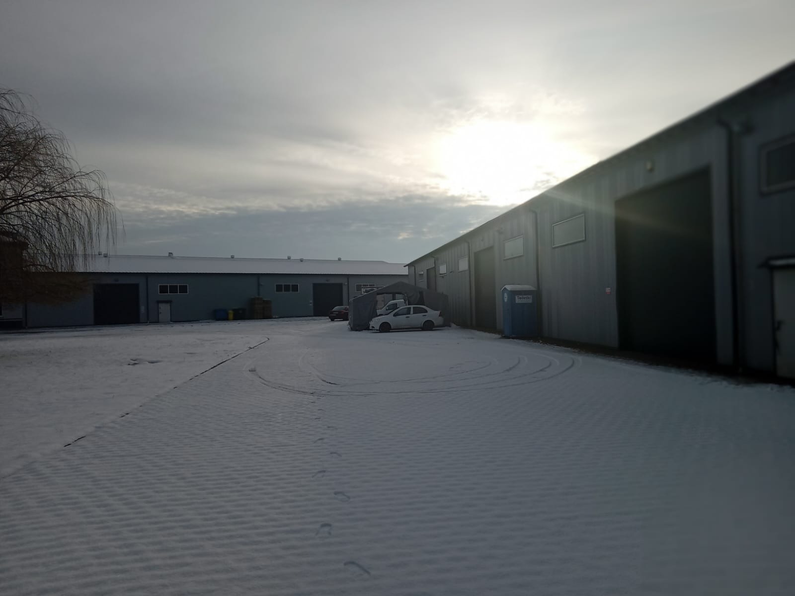 Zimowy widok na teren przemysłowy w Raszynie z halami magazynowymi, pokryty śniegiem z widocznymi śladami opon i pieszych, z przenośną toaletą przy jednej z hal.