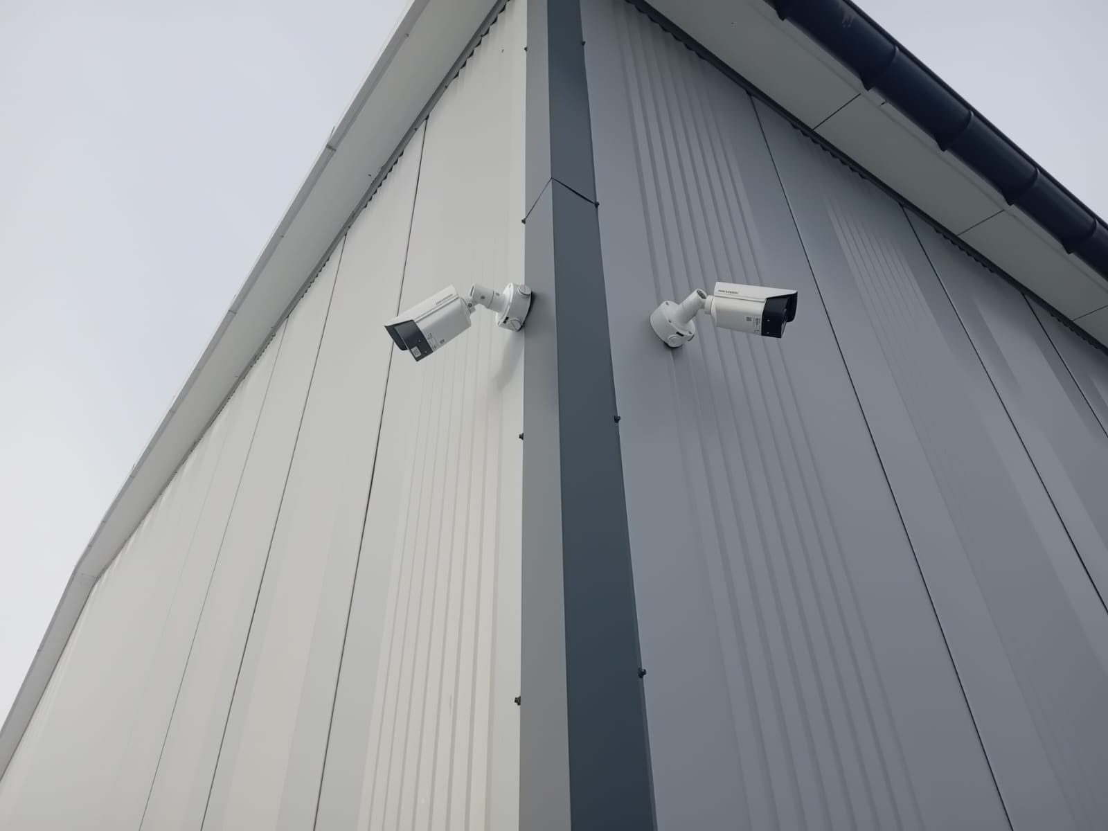 Dwie białe kamery przemysłowe zamontowane na rogu szarego budynku z blachy trapezowej, skierowane w różnych kierunkach.