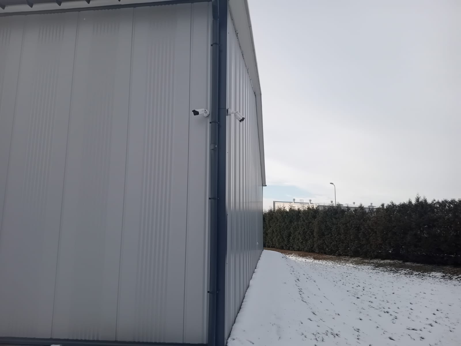 Dwie białe kamery przemysłowe zamontowane na szarej, metalowej ścianie budynku przemysłowego w zimowej scenerii.