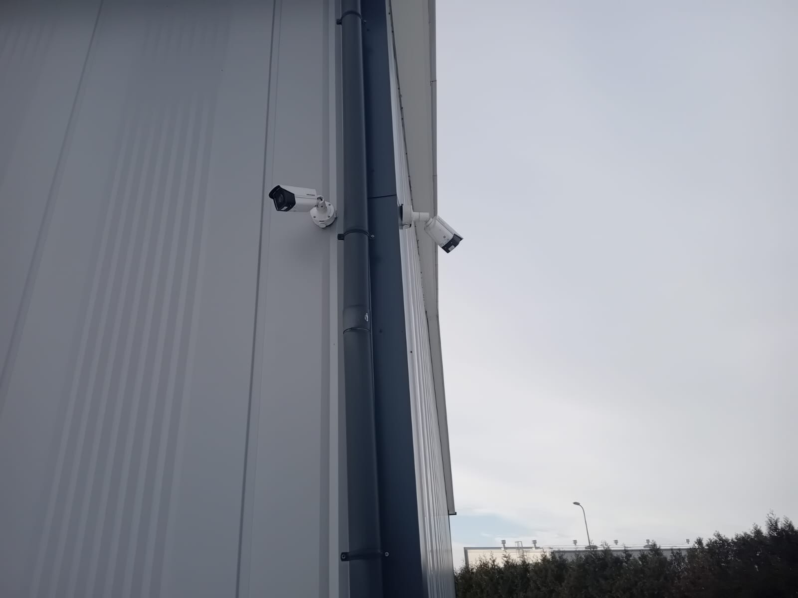Dwie kamery monitoringu zamontowane na zewnętrznej ścianie budynku przemysłowego, widoczne rury spustowe, pochmurne niebo w tle.