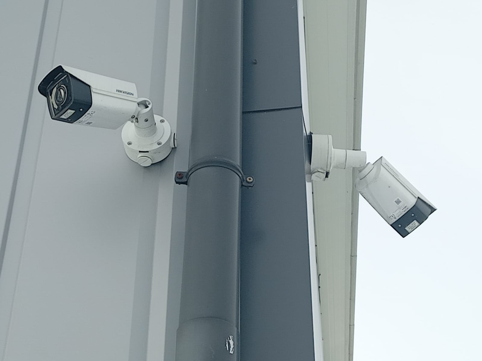 Dwie kamery monitoringu marki Hikvision zamontowane na elewacji budynku, jedna nad rurą spustową, druga pod kątem, skierowana w dół.