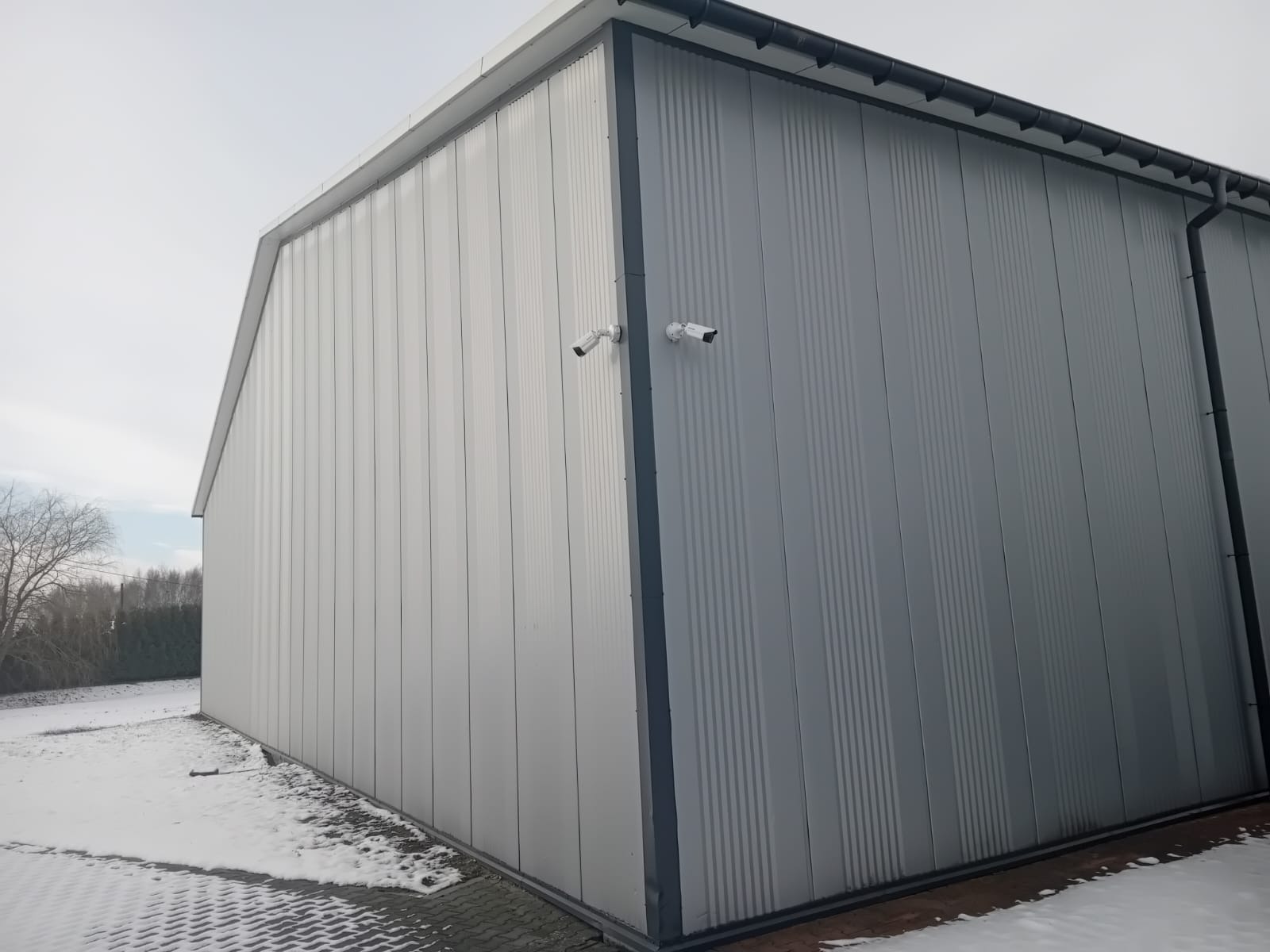 Dwie kamery monitoringu zamontowane na szarej, panelowej ścianie magazynu w zimowej scenerii.