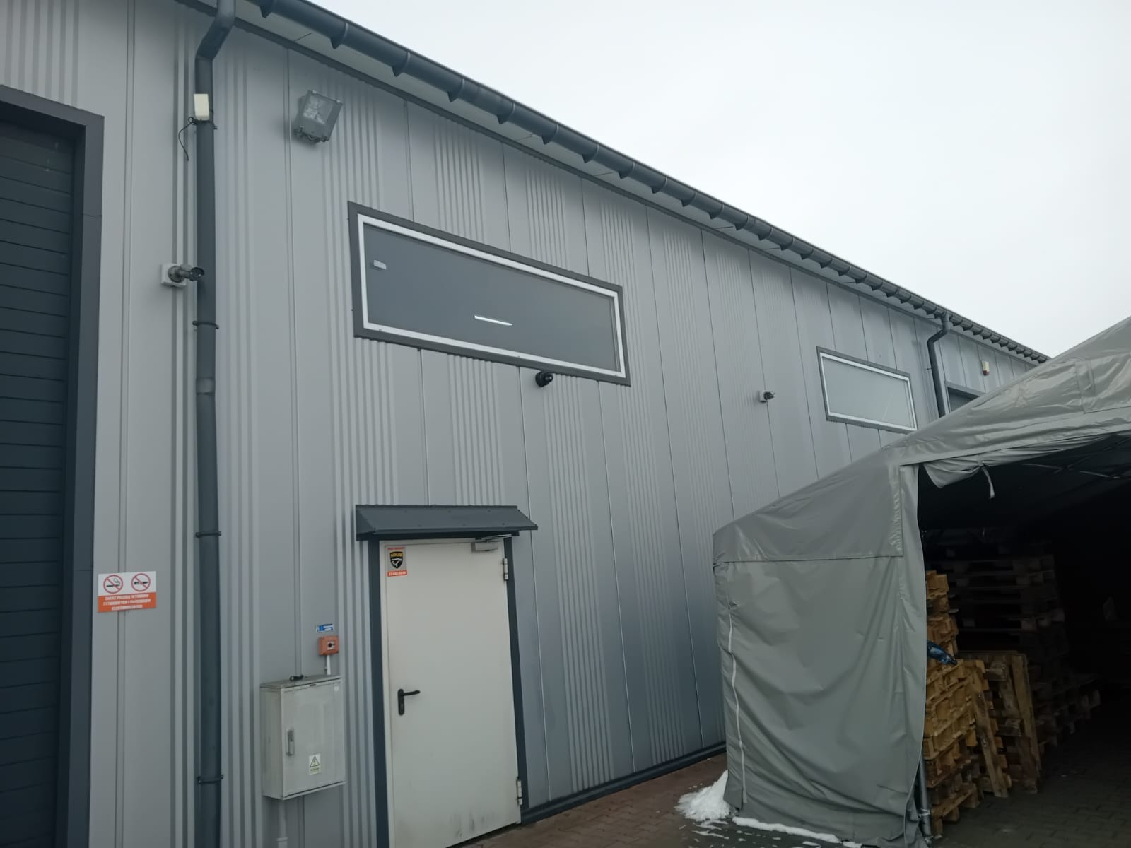 Zewnętrzny widok szarego budynku przemysłowego z zamontowanymi kamerami monitoringu, oknami i drzwiami z systemem alarmowym, częściowo zasłoniętego przez szarą plandekę.