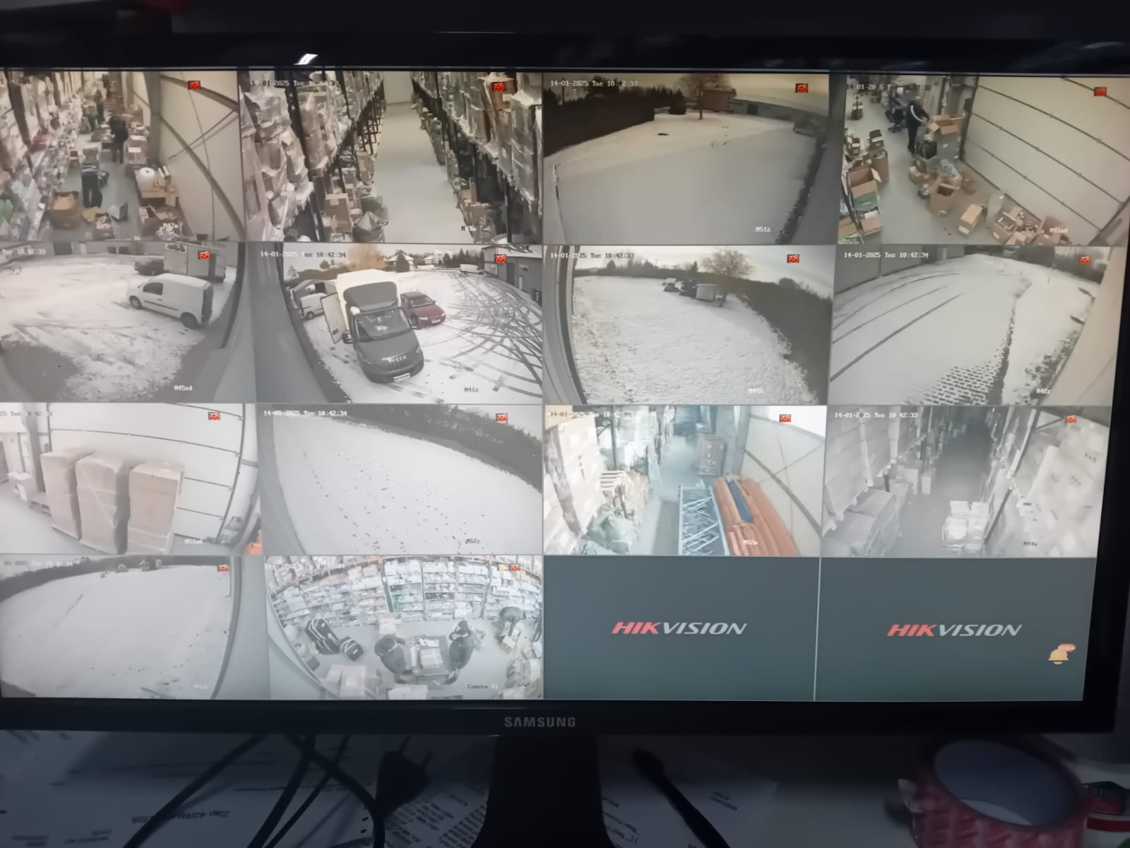 Ekran monitora z widokiem z wielu kamer przemysłowych Hikvision, prezentujących magazyn, parking pokryty śniegiem oraz wnętrze sklepu z klientami.