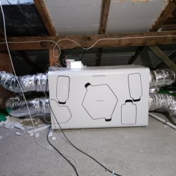 Tomasz Wróblewski - Zamontowany rekuperator Thessla Green z podłączonymi przewodami wentylacyjnymi w przestrzeni poddasza, widoczne drewniane elementy konstrukcyjne dachu i fragmenty izolacji.