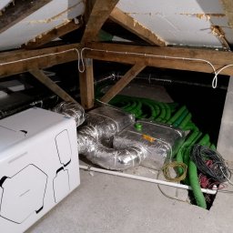 Tomasz Wróblewski - Biała jednostka centralna rekuperatora w surowym pomieszczeniu poddasza, widoczne izolowane kanały wentylacyjne, zielone rury oraz wiązki kabli wychodzące z otworu w podłodze.