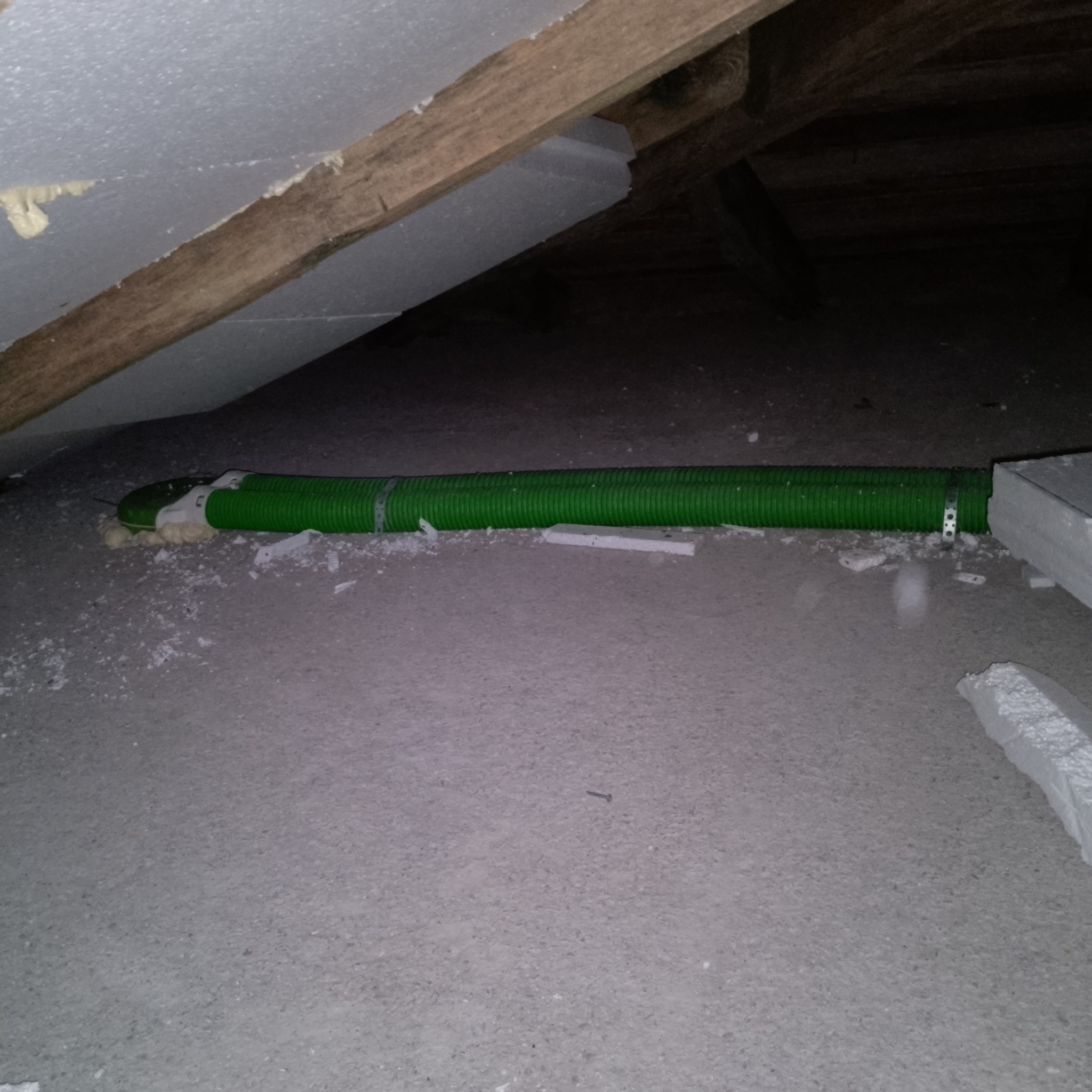 Zielony przewód wentylacyjny, zabezpieczony metalową obejmą, ułożony na betonowej podłodze pod drewnianą konstrukcją dachu, uszczelniony pianką montażową i otoczony fragmentami styropianu.