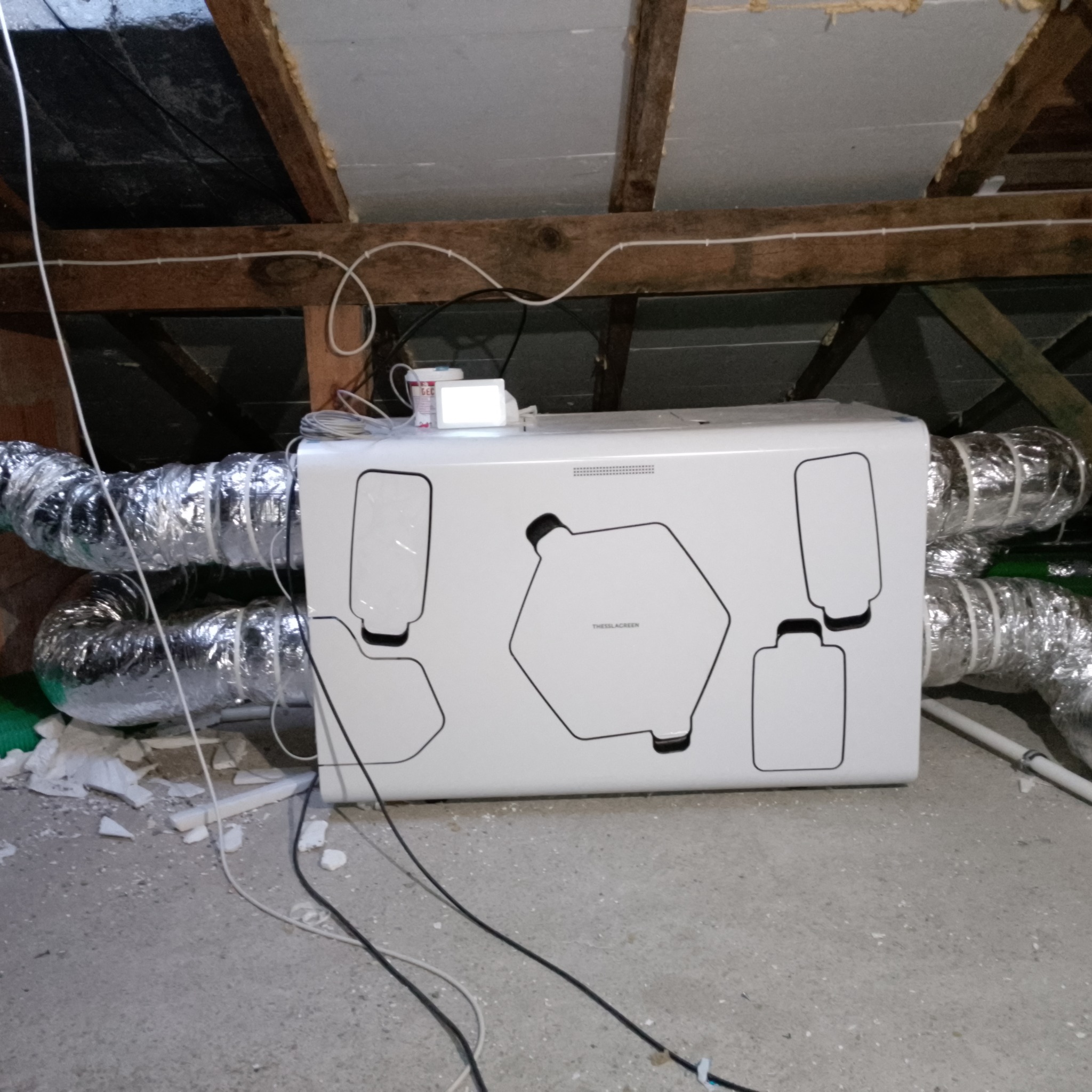 Zamontowany rekuperator Thessla Green z podłączonymi przewodami wentylacyjnymi w przestrzeni poddasza, widoczne drewniane elementy konstrukcyjne dachu i fragmenty izolacji.