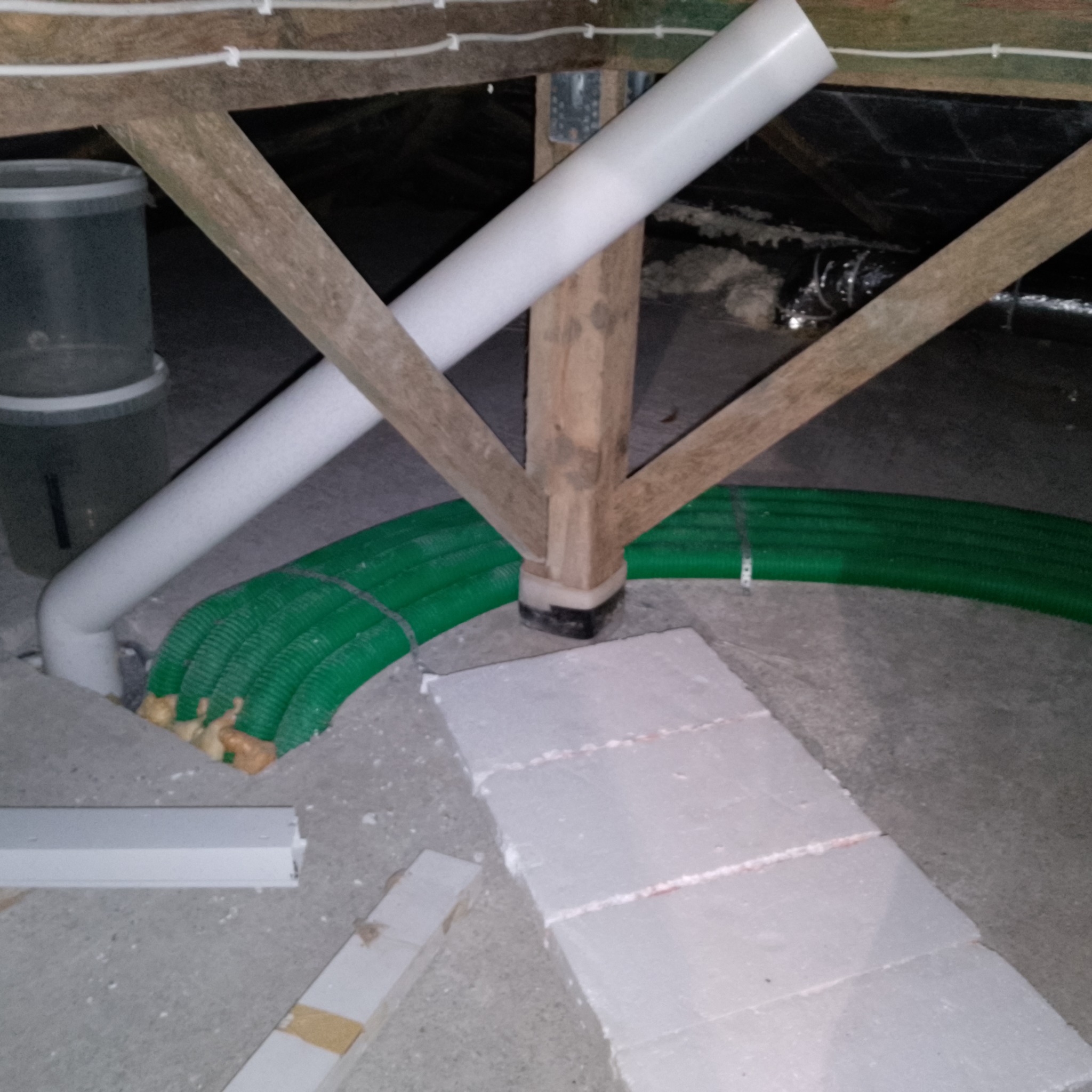 Biała rura wentylacyjna pod skosem, zielone karbowane przewody wentylacyjne ułożone równolegle, drewniana konstrukcja wspierająca, fragment izolacji podłogi.