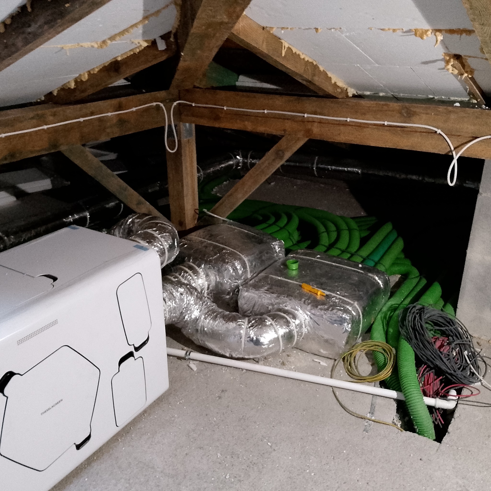 Biała jednostka centralna rekuperatora w surowym pomieszczeniu poddasza, widoczne izolowane kanały wentylacyjne, zielone rury oraz wiązki kabli wychodzące z otworu w podłodze.