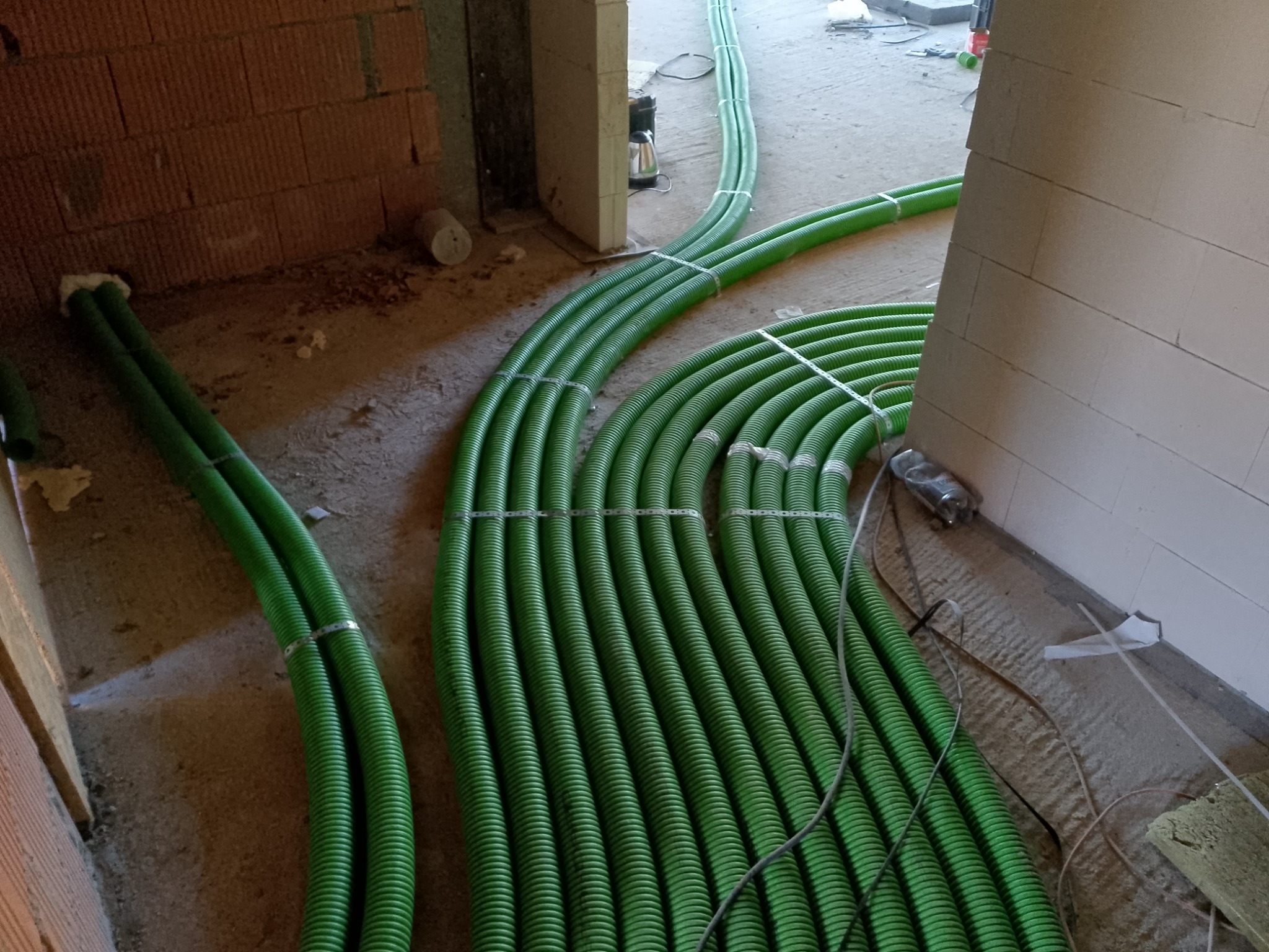 Wiązki zielonych, karbowanych przewodów wentylacyjnych prowadzone w surowym stanie budynku, zabezpieczone metalowymi opaskami.