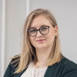 Portret uśmiechniętej blondynki w okularach i ciemnozielonej marynarce, z delikatną biżuterią i subtelnym makijażem na tle jasnej ściany.