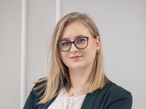 Portret uśmiechniętej blondynki w okularach i ciemnozielonej marynarce, z delikatną biżuterią i subtelnym makijażem na tle jasnej ściany.