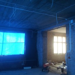 Oliver Cvetković OC-elektryk - Surowy stan wnętrza w Gdańsku podczas remontu, widoczne kable elektryczne prowadzone po suficie i ścianach, okno zasłonięte niebieską folią, w tle nagrzewnica budowlana.