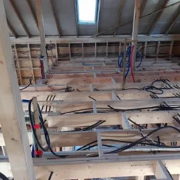 Surowy stan poddasza w trakcie budowy, widoczne liczne przewody elektryczne i rury instalacyjne ułożone między drewnianą konstrukcją podłogi i ścian, oświetlone przez okno dachowe.