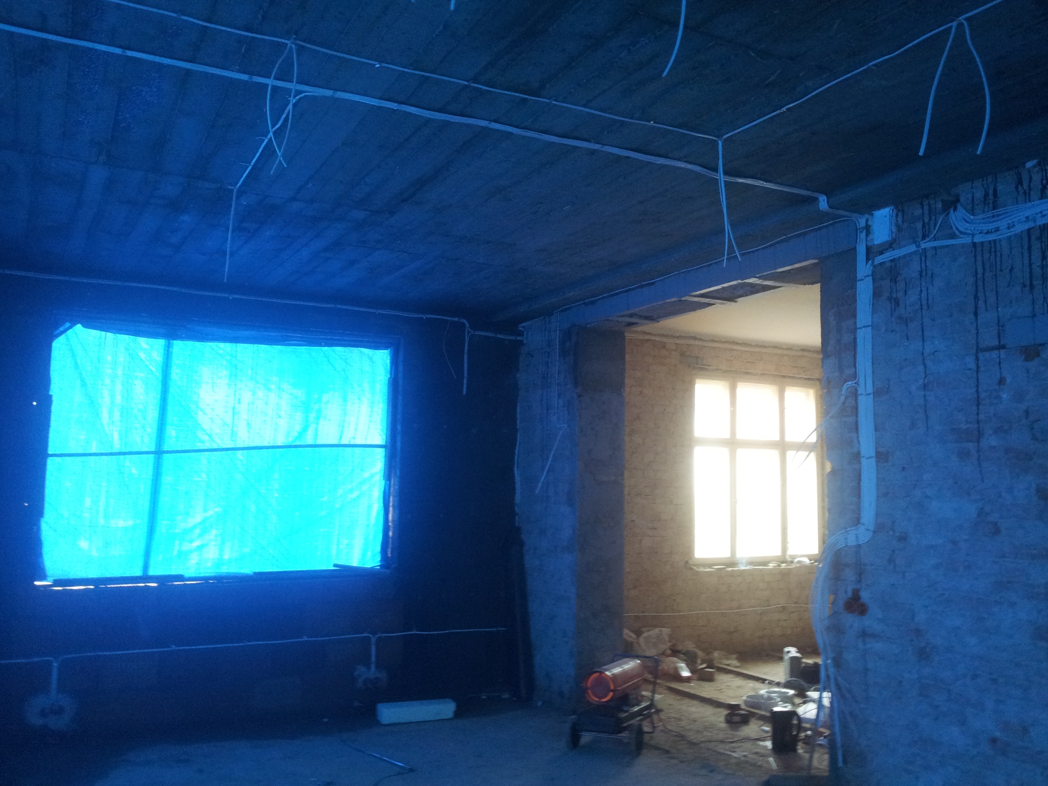 Surowy stan wnętrza w Gdańsku podczas remontu, widoczne kable elektryczne prowadzone po suficie i ścianach, okno zasłonięte niebieską folią, w tle nagrzewnica budowlana.