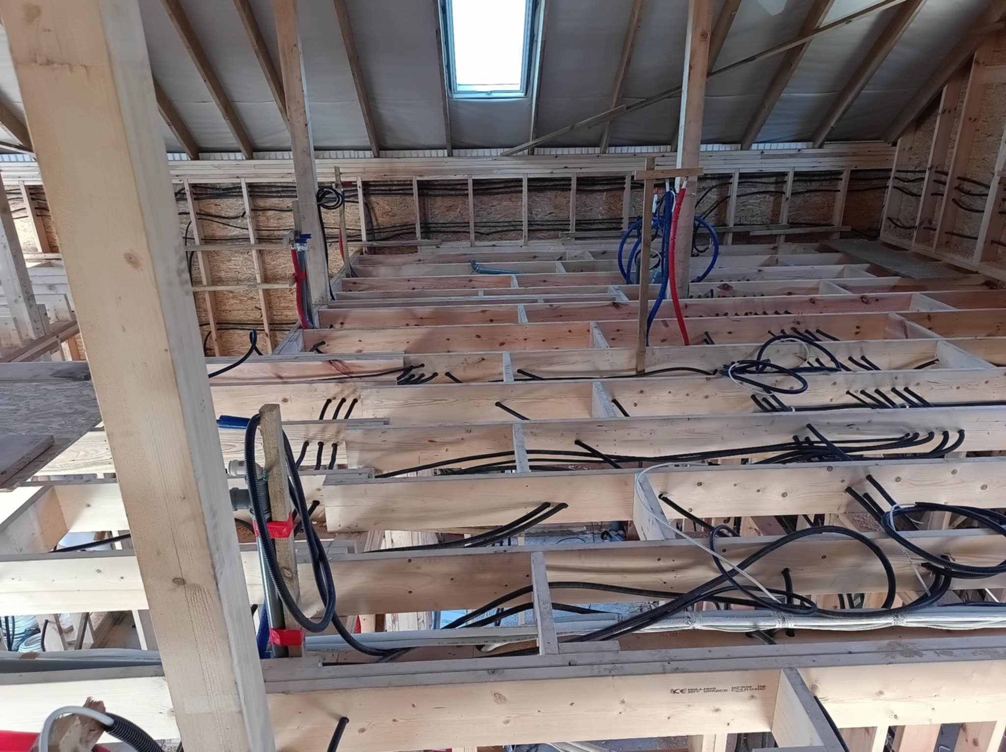 Surowy stan poddasza w trakcie budowy, widoczne liczne przewody elektryczne i rury instalacyjne ułożone między drewnianą konstrukcją podłogi i ścian, oświetlone przez okno dachowe.