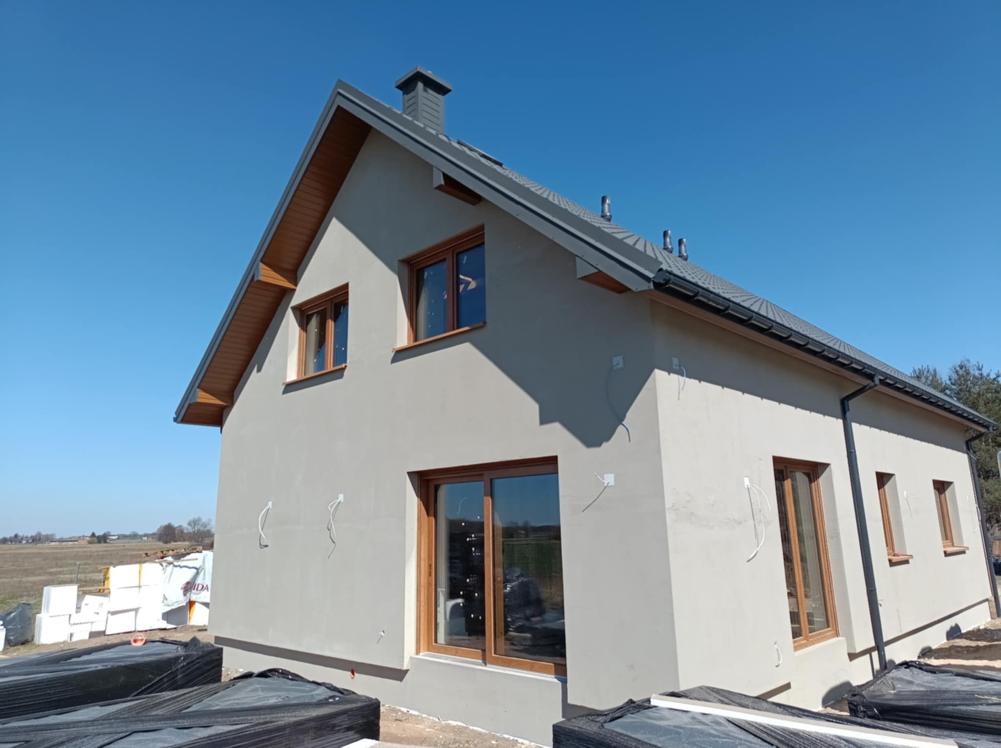 Nowo otynkowany dom jednorodzinny z drewnianymi oknami i wyprowadzonymi przewodami elektrycznymi na zewnątrz budynku, widoczne materiały izolacyjne i budowlane.